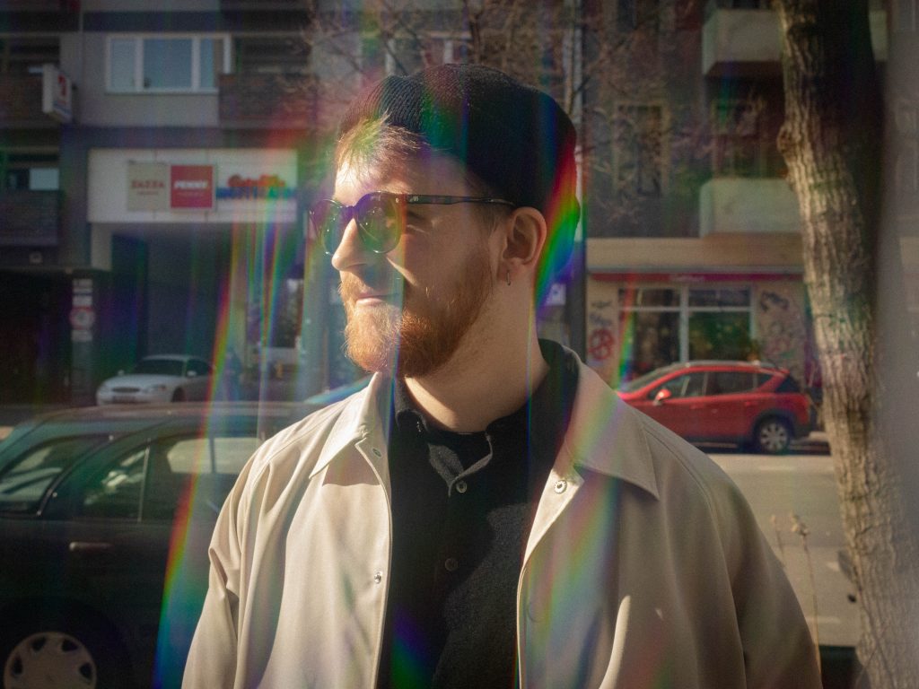 Portrait of Chris Breinl standing in a street looking to the side. He is wearing sunglasses and there raibow coloured lighting effects obstructing the view.
